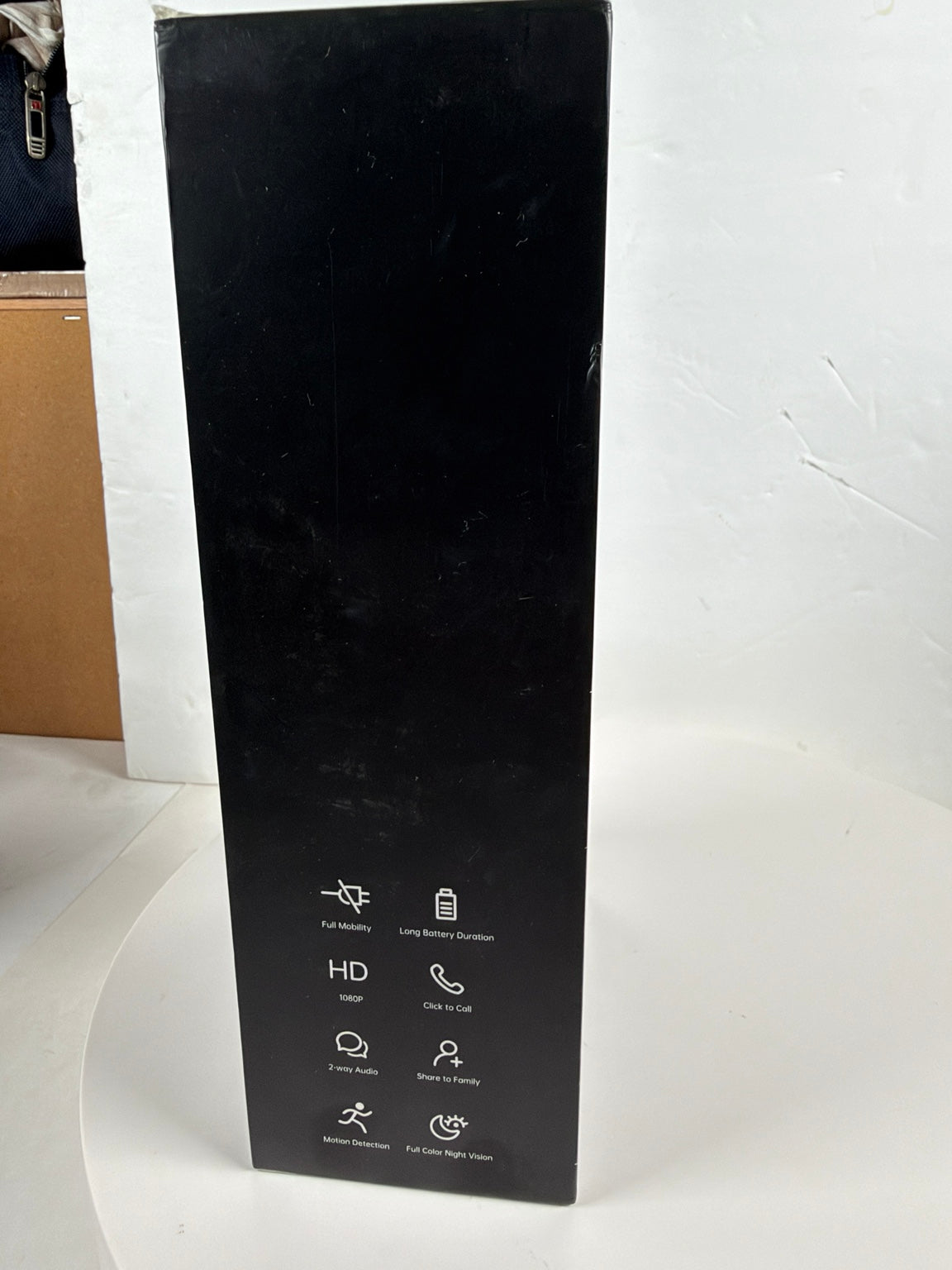 A tall black product box standing upright on a white surface against a white wall, with small white feature icons printed near the bottom.