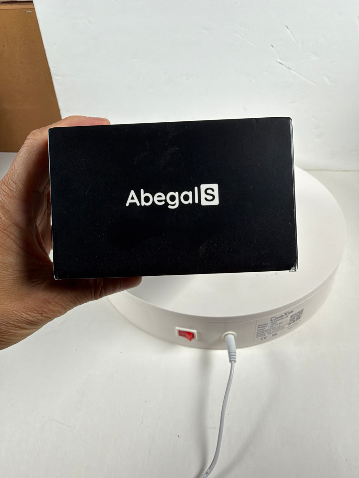 A hand holds a black Abegal S product box in front of a white circular device on a white surface against a white wall.