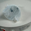 A white plastic replacement part, possibly a filter or cap, wrapped in a clear protective plastic sleeve, resting on a white circular surface. The brand 'CamXim' is visible on the label.