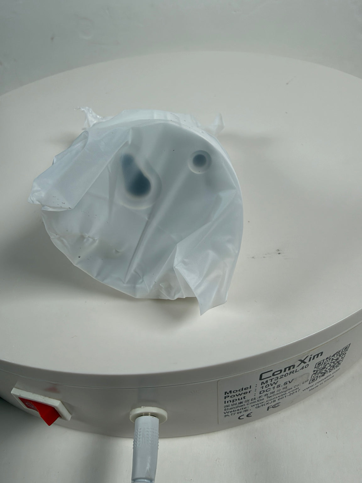 A white plastic replacement part, possibly a filter or cap, wrapped in a clear protective plastic sleeve, resting on a white circular surface. The brand 'CamXim' is visible on the label.