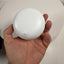 A hand holding a small, round, white plastic object with a small tab-like protrusion on its front, photographed against a white circular backdrop.