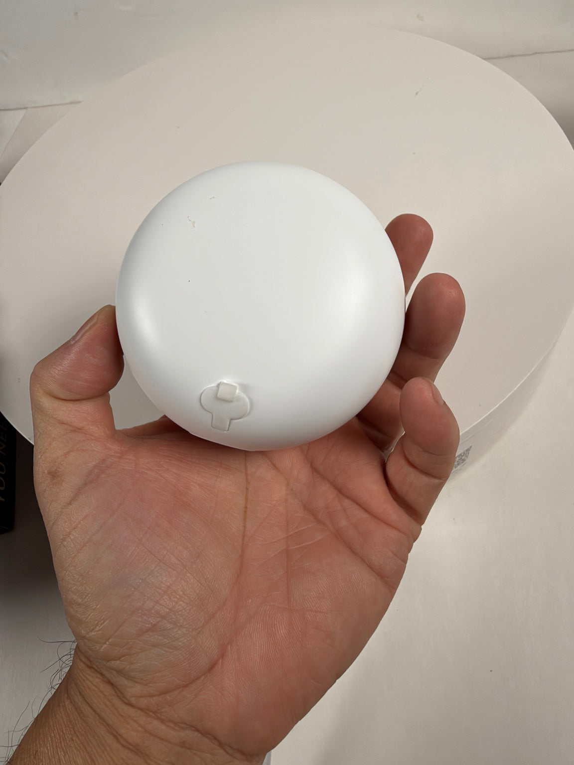 A hand holding a small, round, white plastic object with a small tab-like protrusion on its front, photographed against a white circular backdrop.
