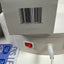 A close-up of a white circular electronic device showing its back panel with a red power switch, a white power cord, a barcode, and a printed box. A blue and white 'Lens Care' sticker is partially visible on the left.