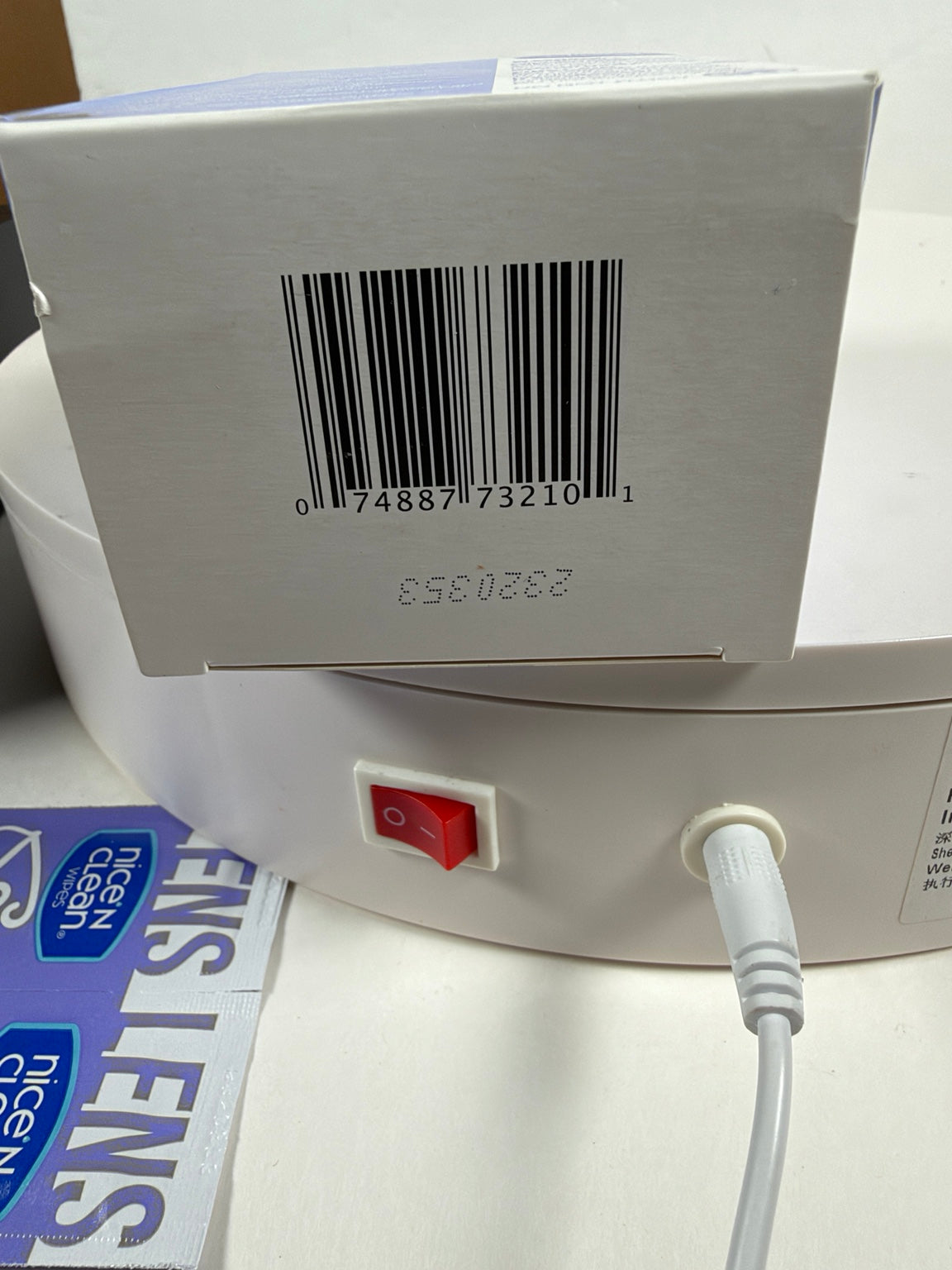 A close-up of a white circular electronic device showing its back panel with a red power switch, a white power cord, a barcode, and a printed box. A blue and white 'Lens Care' sticker is partially visible on the left.