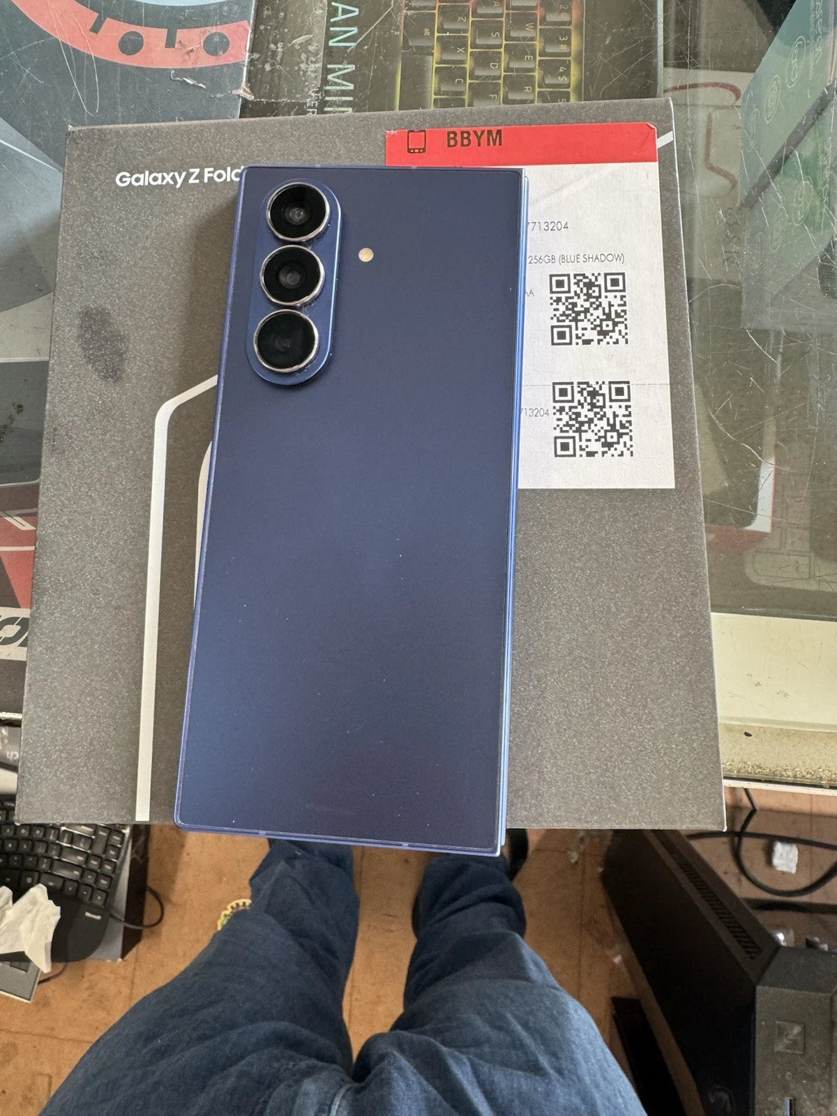 A blue Samsung Galaxy Z Fold smartphone is shown from the back, resting on its original gray packaging box. The box features the Samsung logo and 'Galaxy Z Fold' text.