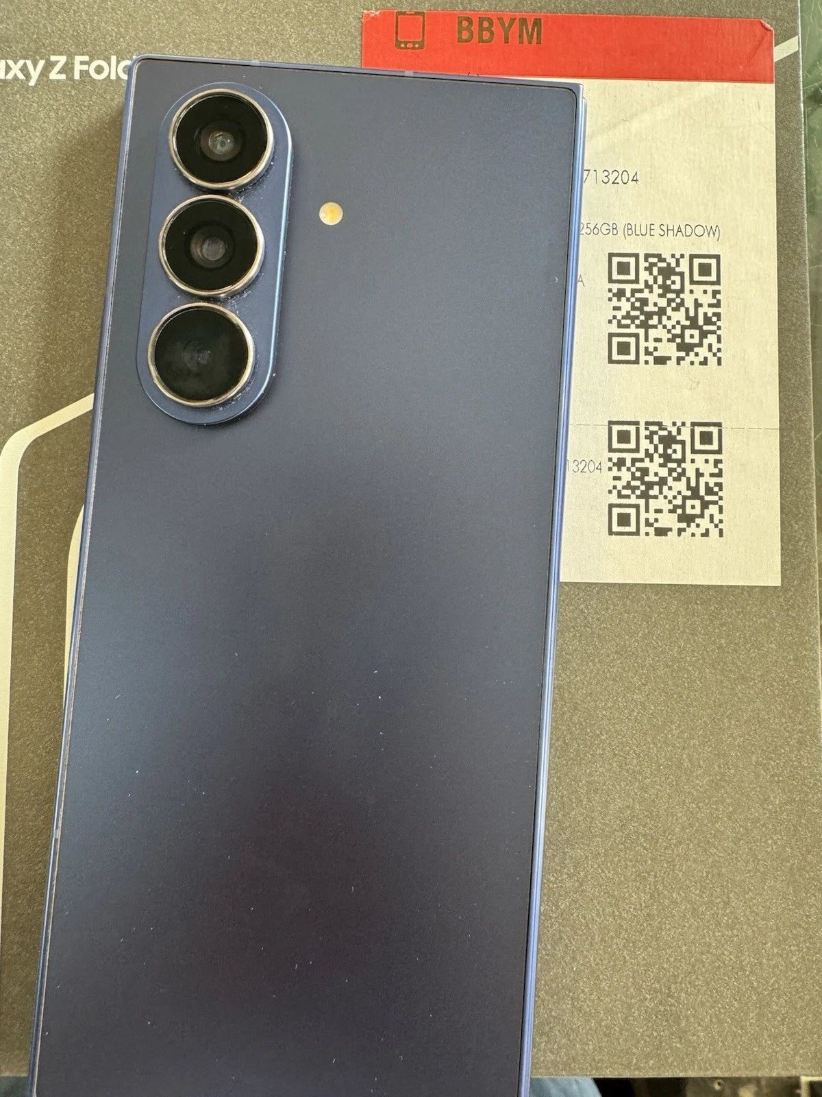 Samsung Galaxy Z Fold 3 smartphone shown from the back in a dark blue-gray color, resting on its original packaging box.