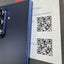 A close-up of a dark blue BBYM smartphone showing its rear camera module and side edge, positioned next to a printed paper tag with QR codes and text reading '7 256GB (BLUE SHADOW)'.