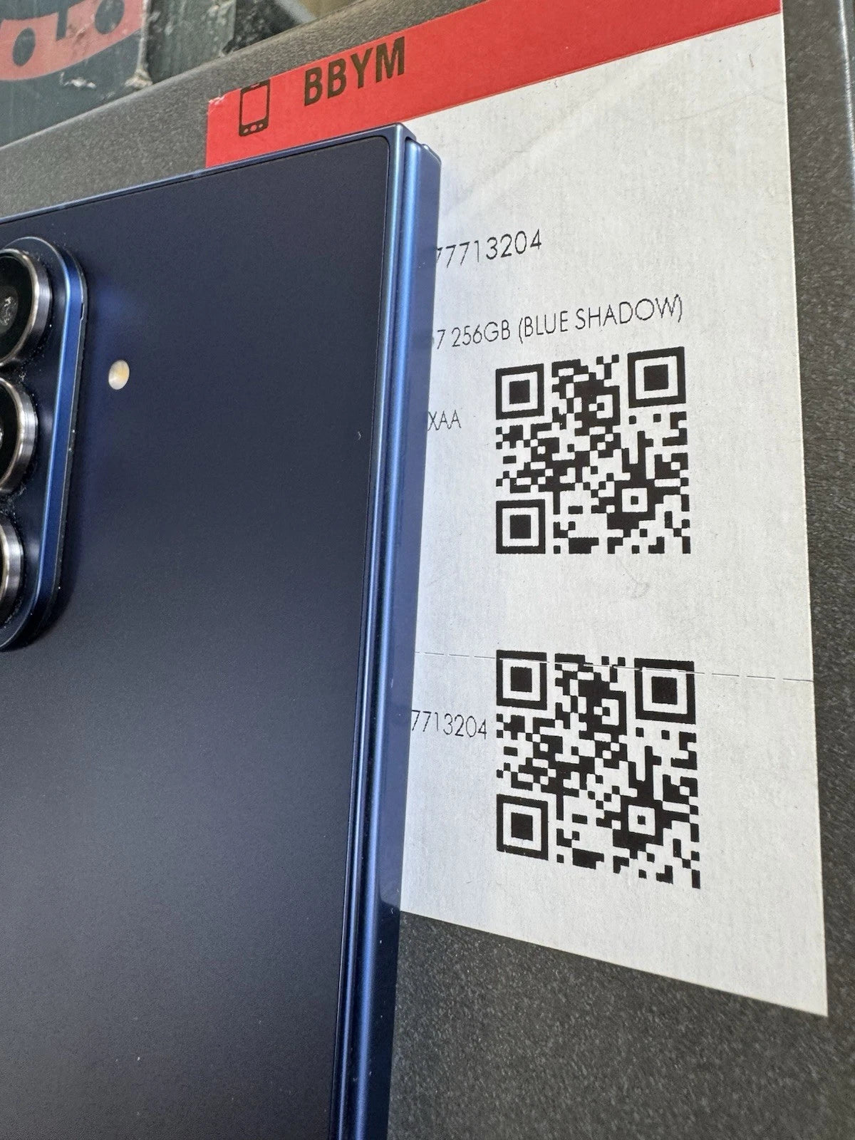 A close-up of a dark blue BBYM smartphone showing its rear camera module and side edge, positioned next to a printed paper tag with QR codes and text reading '7 256GB (BLUE SHADOW)'.