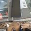 A black Samsung Galaxy smartphone with a blue edge frame rests on top of its original product box. The photo is taken from a first-person perspective, showing the person's legs in blue jeans and black flip-flops on a tiled floor, with a messy desk visible in the background.