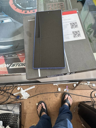 A black Samsung Galaxy smartphone with a blue edge frame rests on top of its original product box. The photo is taken from a first-person perspective, showing the person's legs in blue jeans and black flip-flops on a tiled floor, with a messy desk visible in the background.