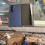 A blue Samsung Galaxy S24 Ultra smartphone shown from the back, resting on a display counter in a retail or repair shop environment. The phone is positioned next to its original packaging box.