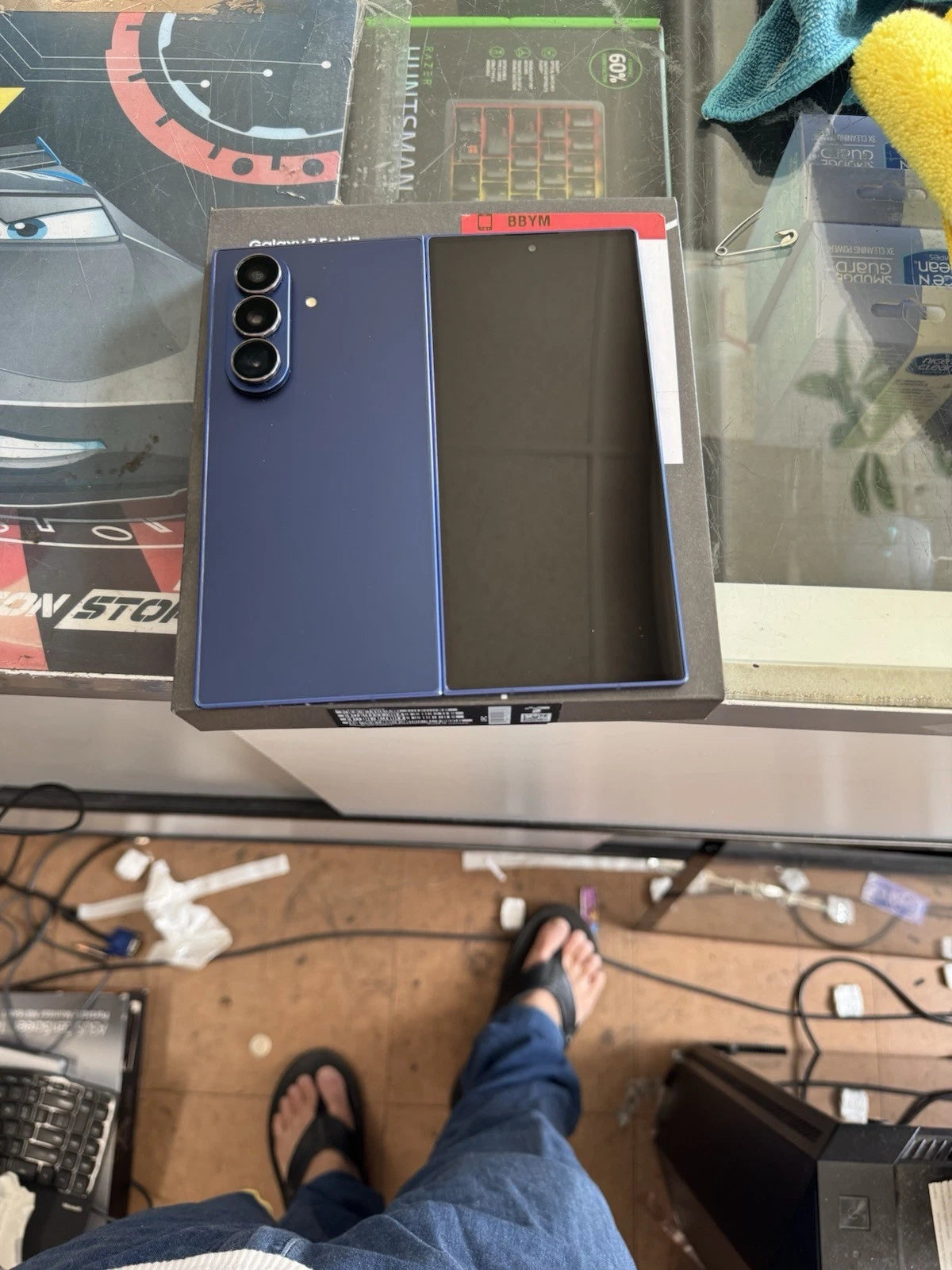 A blue Samsung Galaxy S24 Ultra smartphone shown from the back, resting on a display counter in a retail or repair shop environment. The phone is positioned next to its original packaging box.