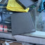A dark gray laptop lid and matching inner box are shown on a glass desk next to a yellow microfiber cloth, with a messy workspace visible underneath.