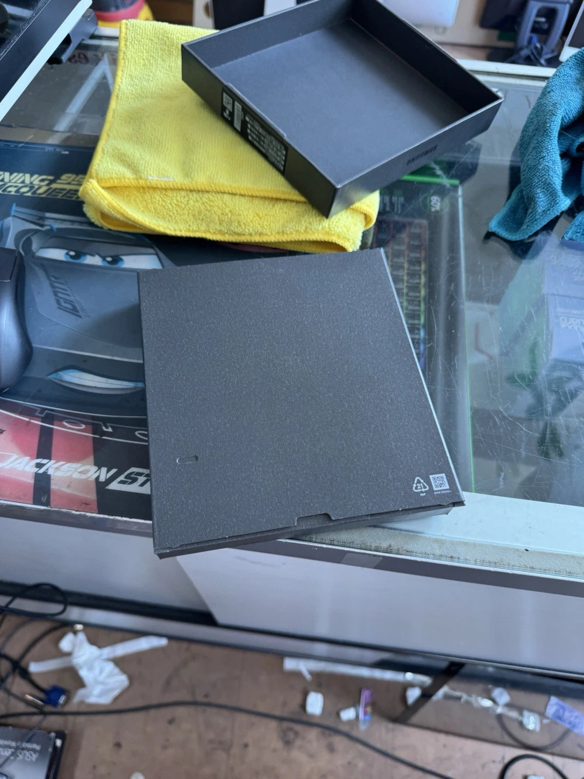 A dark gray laptop lid and matching inner box are shown on a glass desk next to a yellow microfiber cloth, with a messy workspace visible underneath.