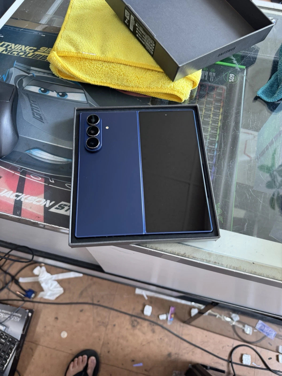A Samsung Galaxy S24 Ultra smartphone in a dark blue and black finish, shown inside its original retail box on a glass desk with a yellow microfiber cloth and accessories.