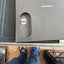 A close-up of a Samsung-branded dark gray textured box with a cutout showing a small white object inside, photographed from above on a desk with a person's legs and feet visible below.
