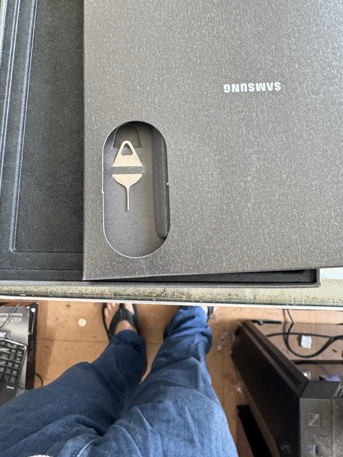 A close-up of a Samsung-branded dark gray textured box with a cutout showing a small white object inside, photographed from above on a desk with a person's legs and feet visible below.