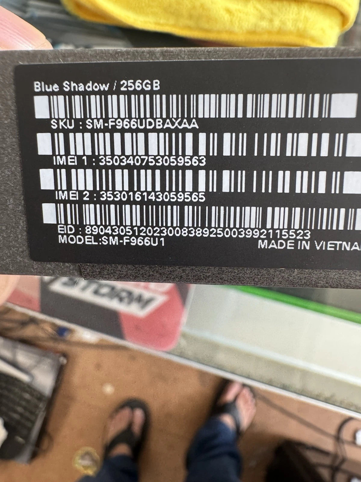 A close-up of a black product label with white text and barcode, showing 'Blue Shadow / 256GB' and product identifiers. A hand is holding the label, and a real indoor environment with floor, sandals, and other objects is visible in the background.