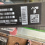 A close-up of a black product label with barcode, UPC, QR code, and 'MADE IN VIETNAM' text, photographed in a real environment with a person's feet and sandals visible below.