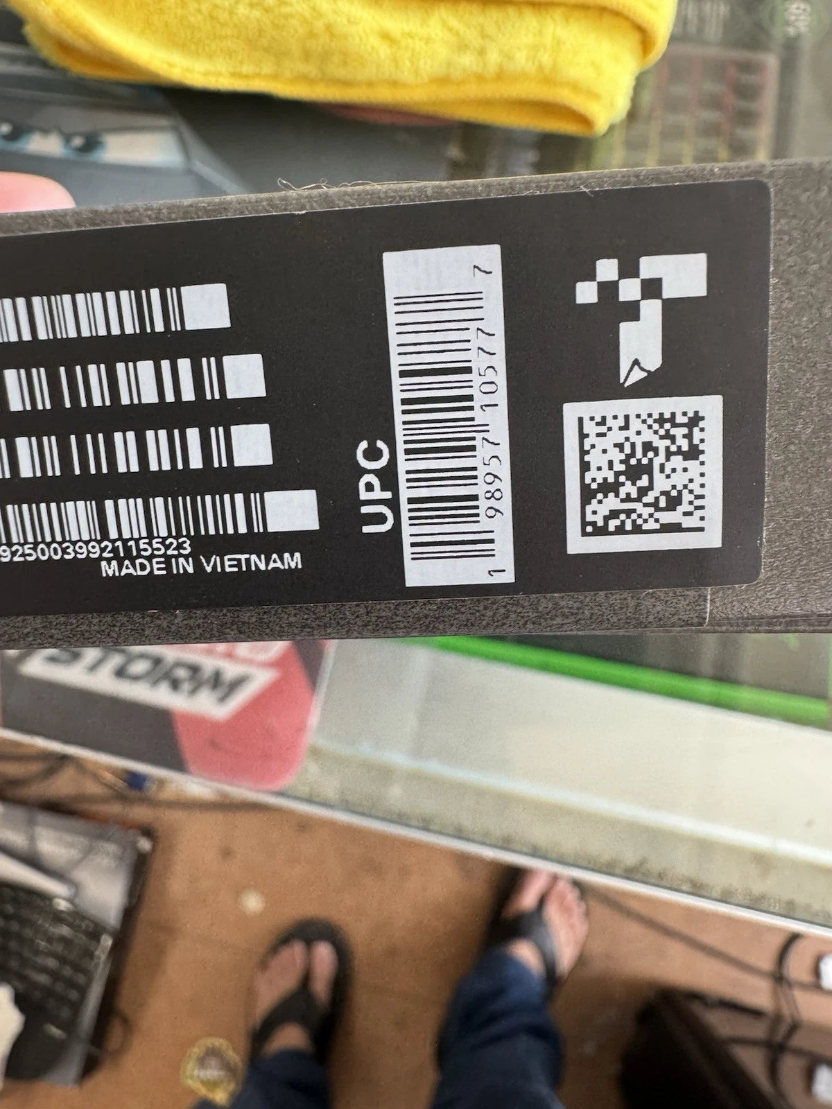 A close-up of a black product label with barcode, UPC, QR code, and 'MADE IN VIETNAM' text, photographed in a real environment with a person's feet and sandals visible below.