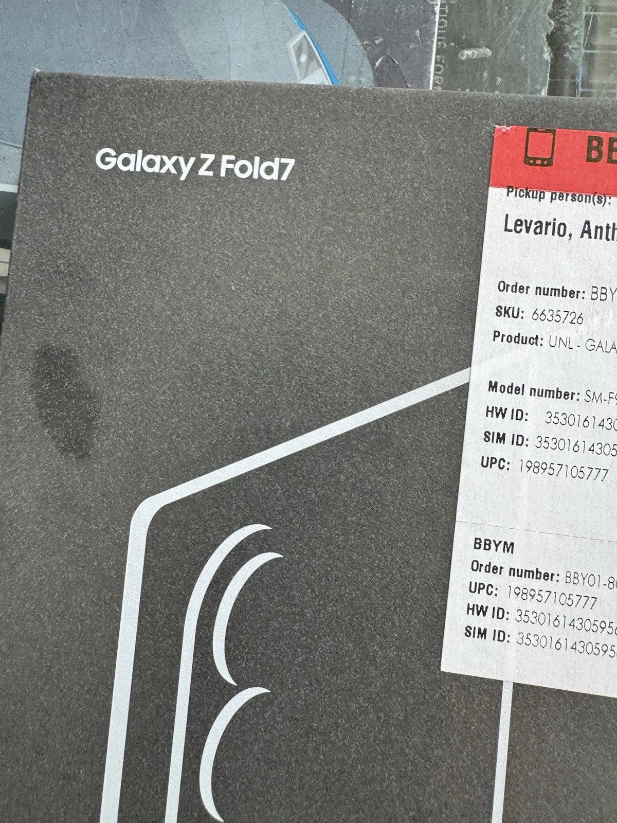 A black Samsung Galaxy Z Fold7 retail box with white branding and curved graphic lines, shown next to a white shipping/pickup label with red header.