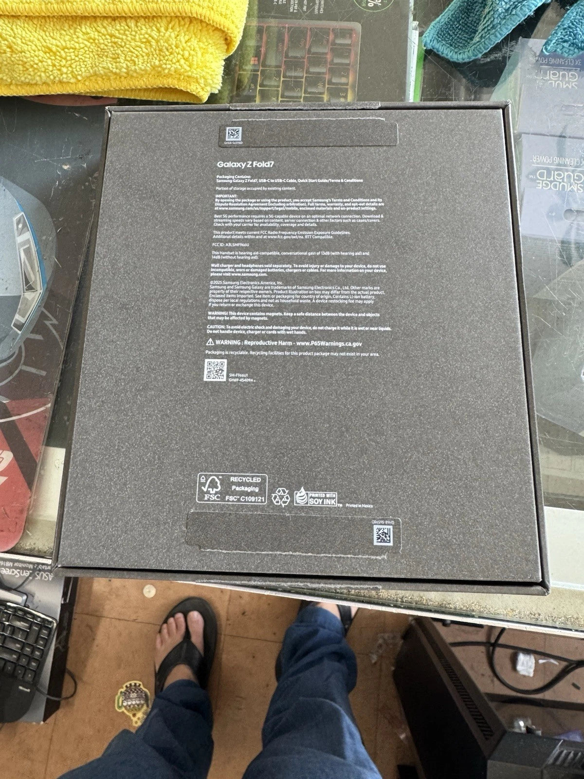 The back of a Samsung Galaxy Z Fold 7 product box, showing printed specifications and icons on a dark gray surface, photographed on a desk with a person's feet visible at the bottom.