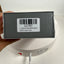 A hand holds a dark gray rectangular product box wrapped in clear plastic, showing a white barcode label with text. The box is positioned above a white circular device with a power cord plugged in against a plain white background.