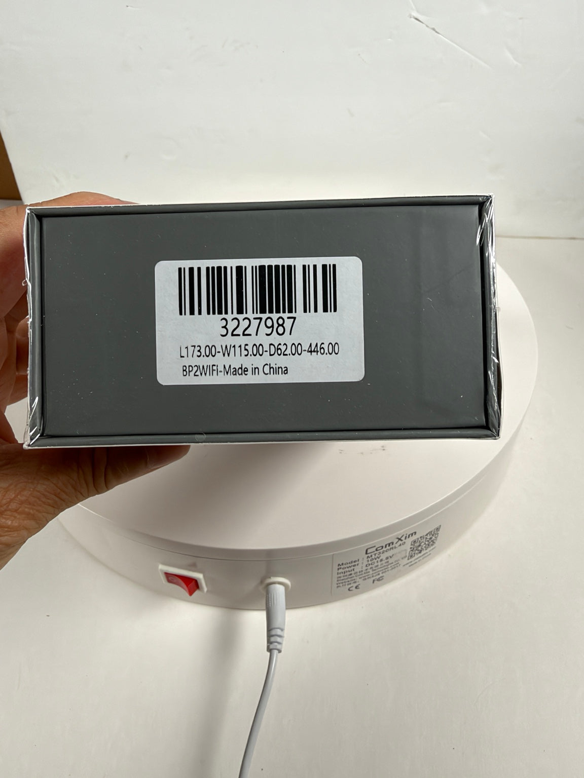 A hand holds a dark gray rectangular product box wrapped in clear plastic, showing a white barcode label with text. The box is positioned above a white circular device with a power cord plugged in against a plain white background.