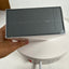 A hand holding a dark gray rectangular product box with a clear plastic top, positioned over a white circular device on a white surface.
