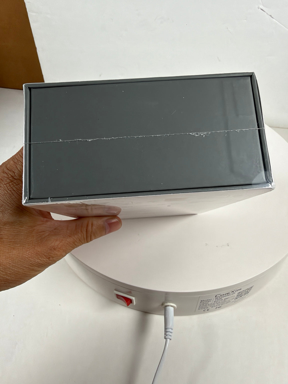 A hand holding a dark gray rectangular product box with a clear plastic top, positioned over a white circular device on a white surface.