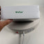 A hand holds a white Wellue product box wrapped in clear plastic, with the green 'Wellue' logo printed on the top. Below the box, a round white device with a power cord is visible against a plain light background.