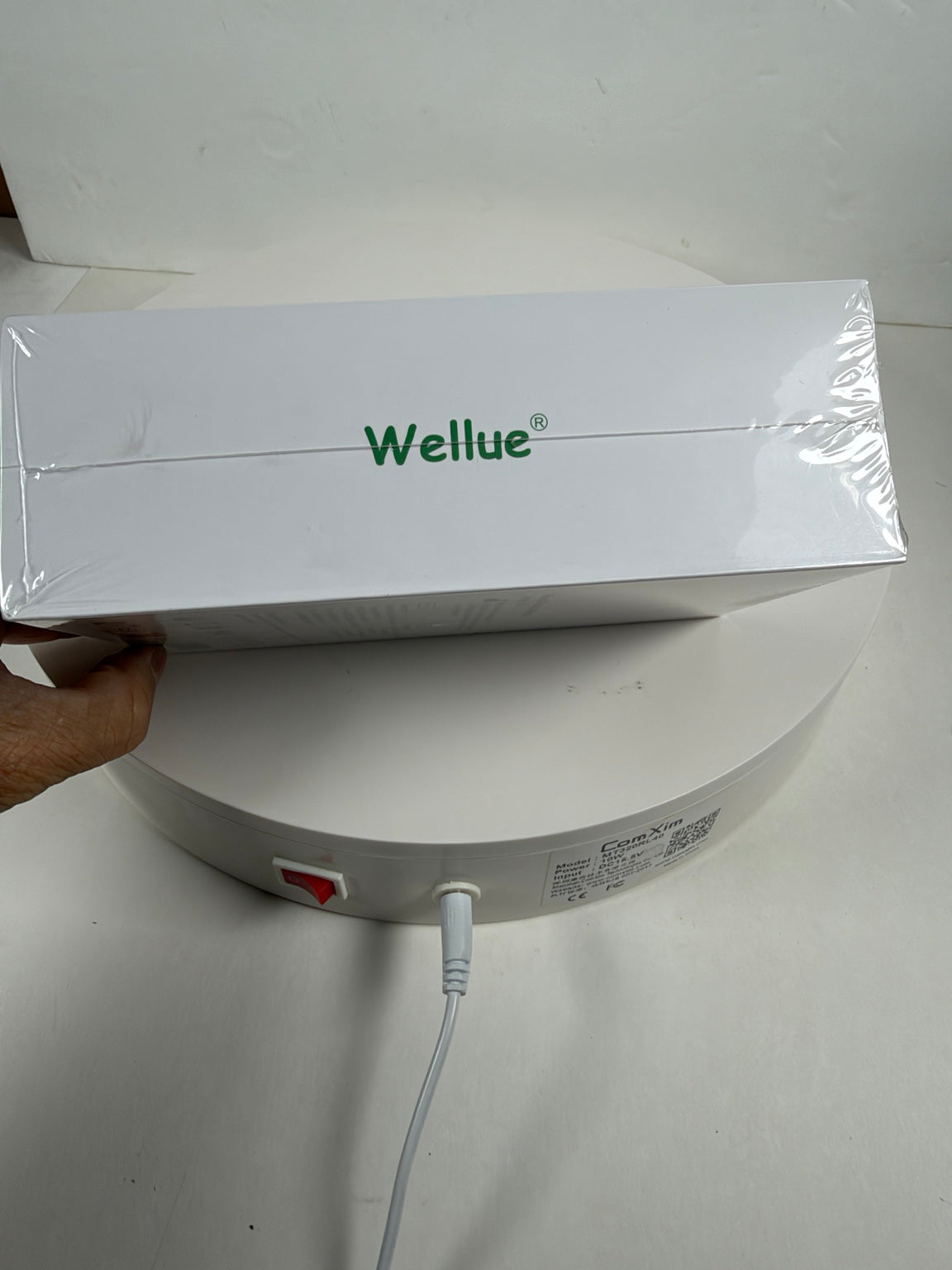 A white Wellue product box wrapped in clear plastic, held by a hand over a white round base with a power cord plugged in.