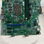 A green computer motherboard with an LGA socket, two black RAM slots, and various electronic components, photographed from above on a white circular platform.