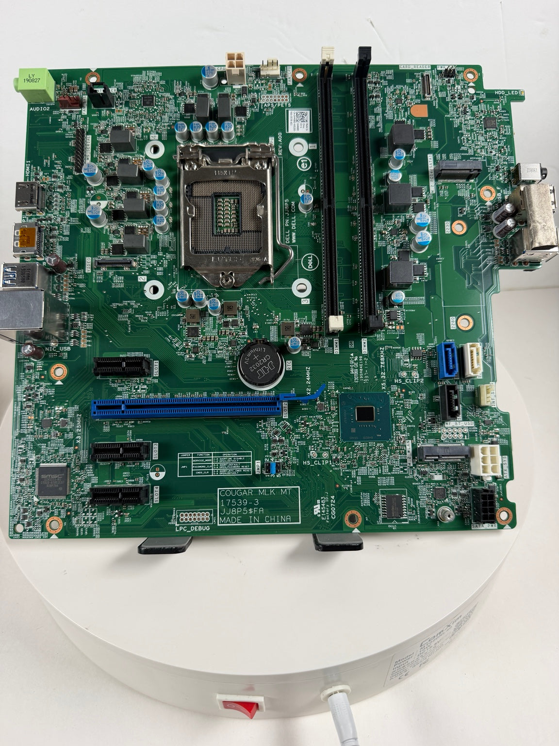 A green computer motherboard with an LGA socket, two black RAM slots, and various electronic components, photographed from above on a white circular platform.
