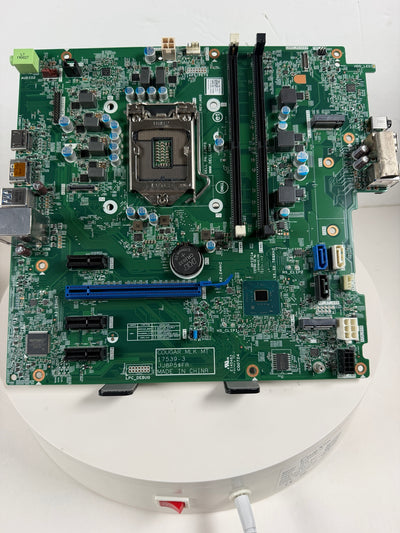 A green computer motherboard with an LGA socket, two black RAM slots, and various electronic components, photographed from above on a white circular platform.