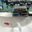 Close-up of a white cylindrical electronic device with a red power switch and a power cord plugged into it, showing a green circuit board with USB ports and a metal mesh component.