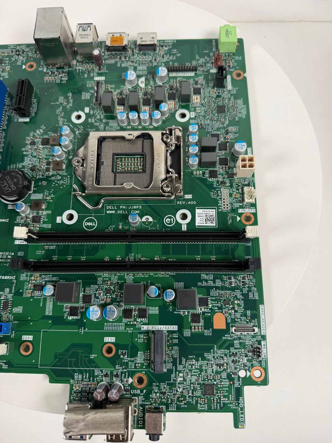 A green Dell motherboard with an LGA 1200 CPU socket, RAM slots, and various electronic components, photographed from a top-down angle on a white surface.