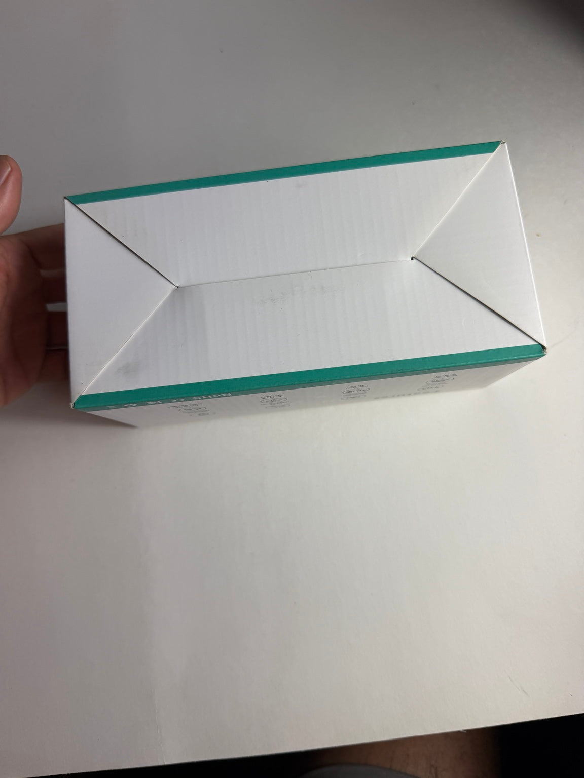 A white rectangular product box with teal-green edges, held by a hand on a plain white surface.