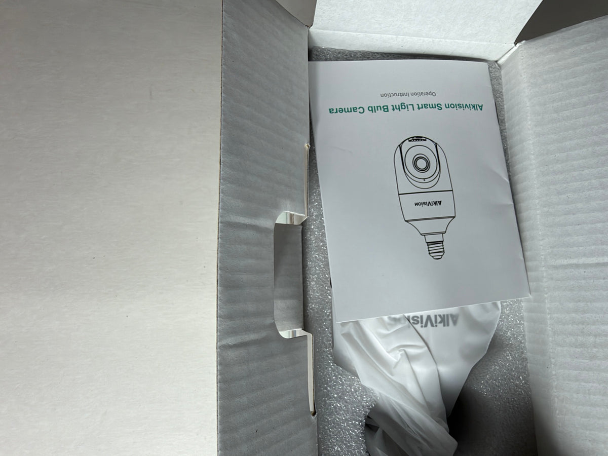 An open cardboard box containing a white paper manual for the Aqvidis Alibaba Smart Light Bulb Camera, nestled in gray protective foam.