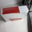 A rectangular product box wrapped in clear plastic, featuring a bold geometric color-blocked design in red, white, and a muted pinkish-red, photographed on a white surface.