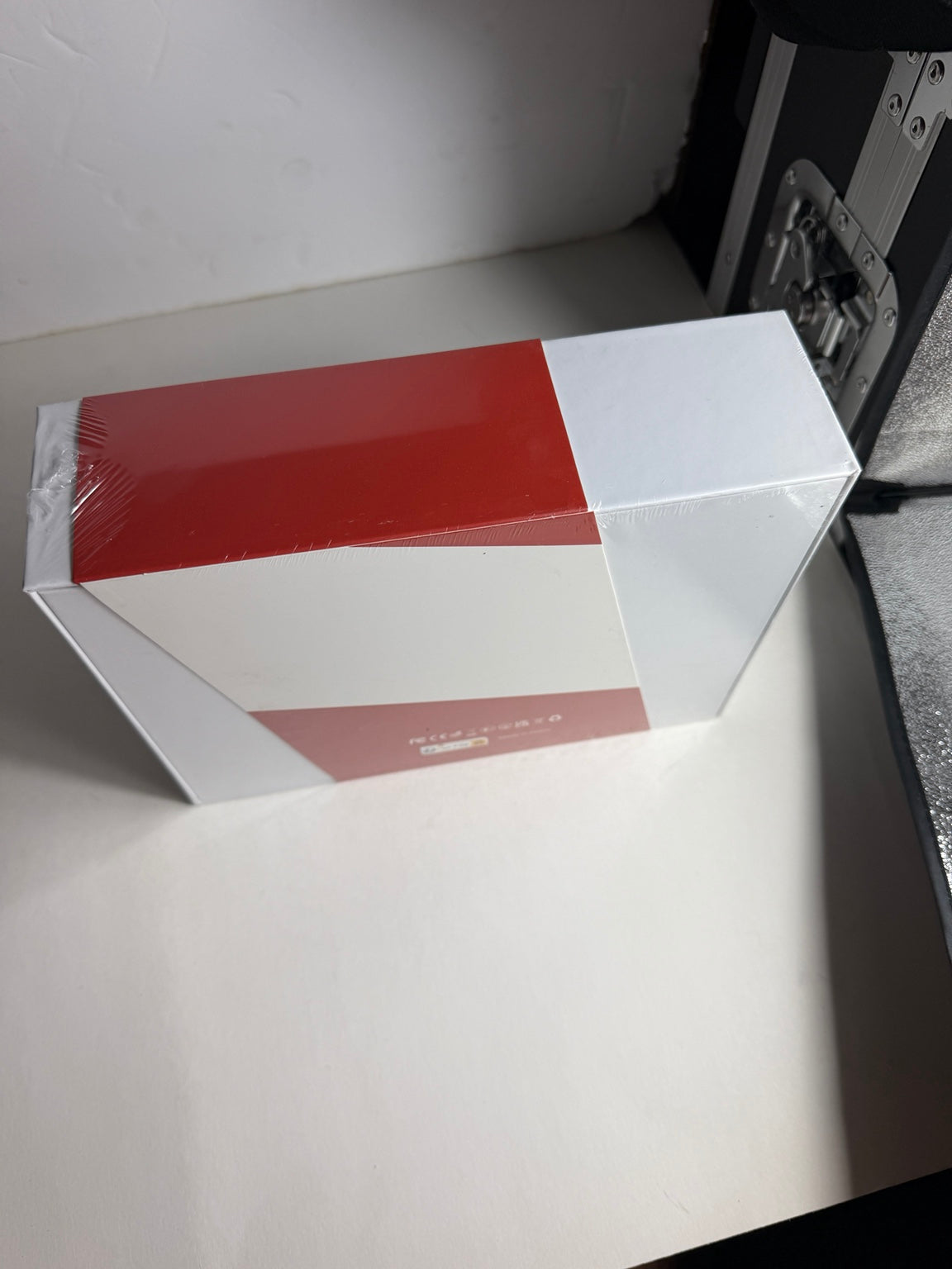 A rectangular product box wrapped in clear plastic, featuring a bold geometric color-blocked design in red, white, and a muted pinkish-red, photographed on a white surface.