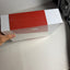 A hand holding a rectangular product box with a red top and white sides, photographed on a white surface against a white wall.