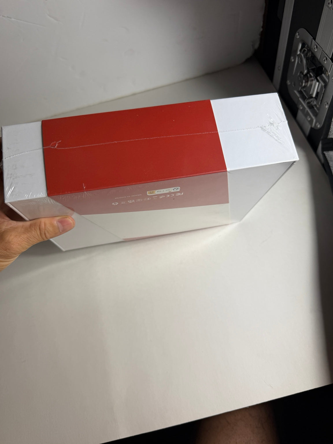 A hand holding a rectangular product box with a red top and white sides, photographed on a white surface against a white wall.