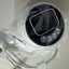 A round black and white electronic device wrapped in clear plastic packaging, with attached white cables and a green connector, photographed on a light neutral surface.