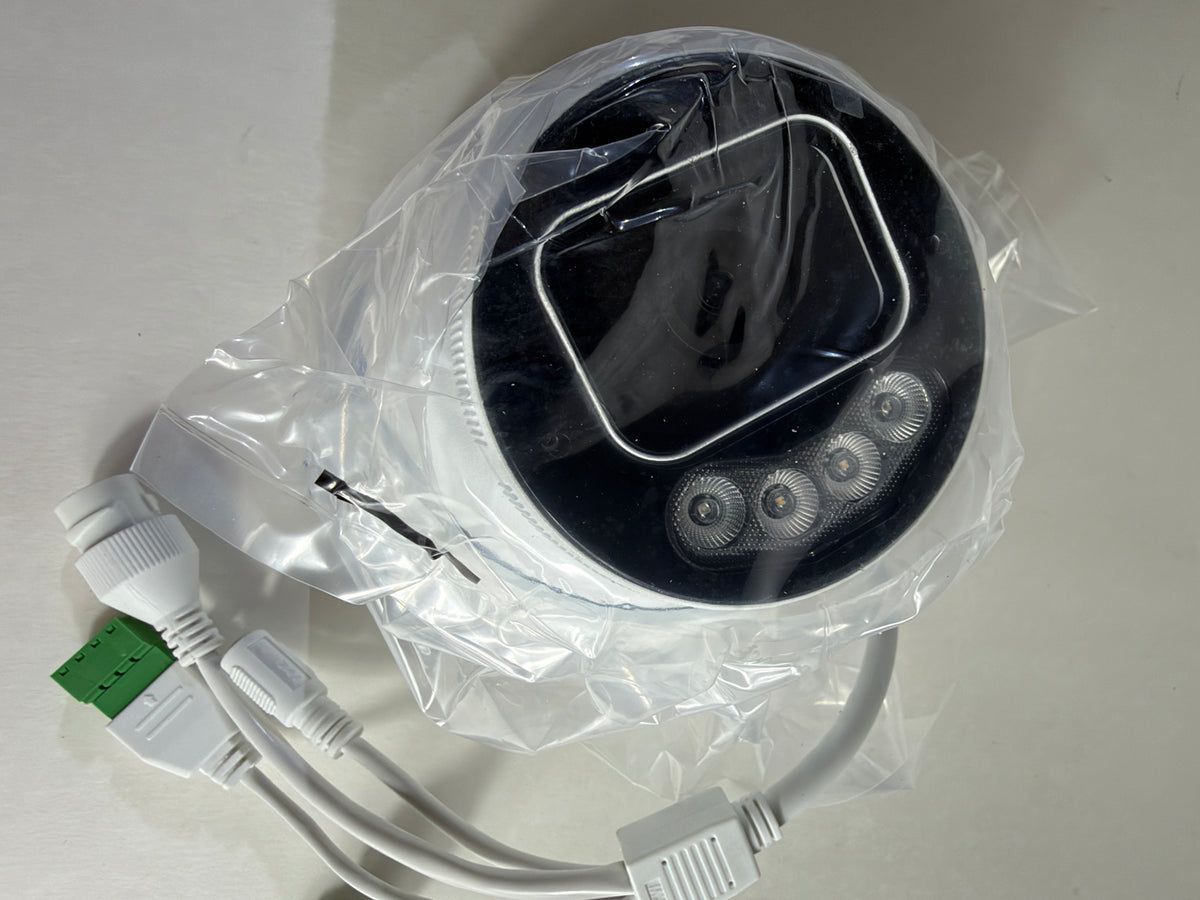 A round black and white electronic device wrapped in clear plastic packaging, with attached white cables and a green connector, photographed on a light neutral surface.
