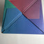 A square box with a geometric, folded triangular design in multiple colors including blue, purple, red, and green, photographed on a plain off-white background.