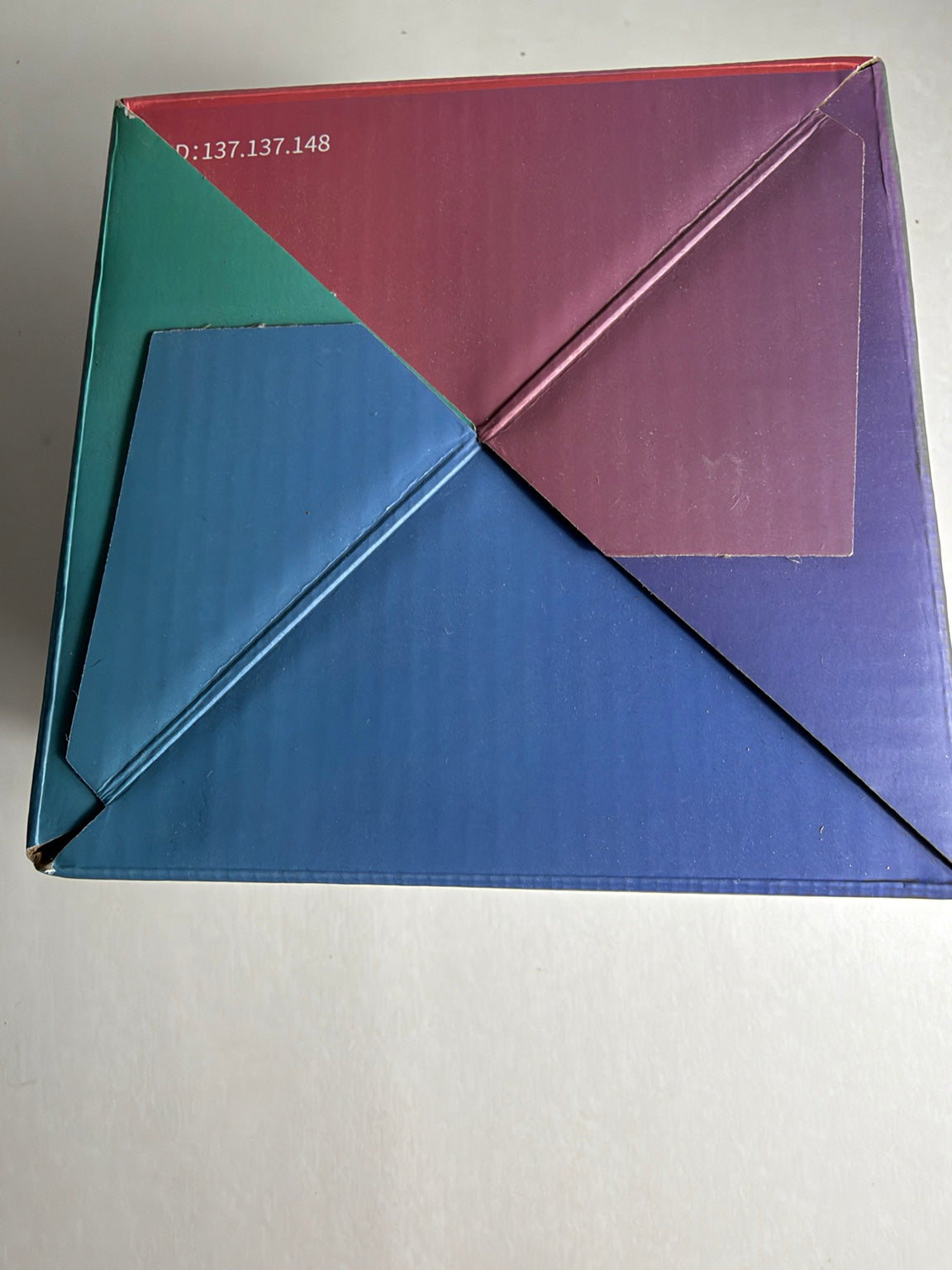 A square box with a geometric, folded triangular design in multiple colors including blue, purple, red, and green, photographed on a plain off-white background.