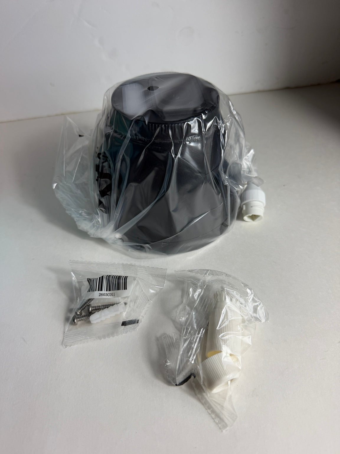 A black cylindrical appliance part, likely a pump or filter component, wrapped in clear plastic packaging, with two small accessory bags containing white fittings and metal pieces on a white surface.