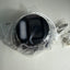 A black circular camera or sensor device wrapped in clear plastic packaging, with a white cable visible on a light gray surface.