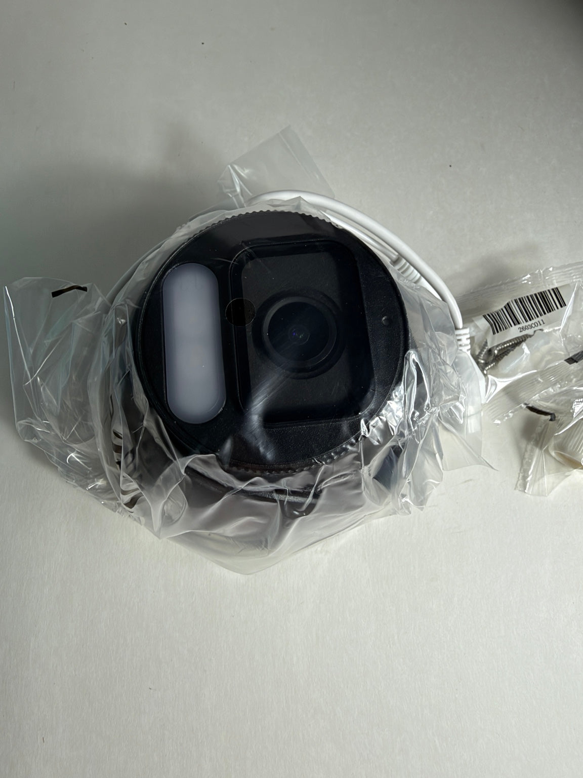 A black circular camera or sensor device wrapped in clear plastic packaging, with a white cable visible on a light gray surface.